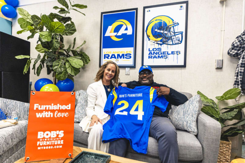 Two people sitting on a gray couch holding a blue sports jersey with "BOB'S FURNITURE" and number "24" in yellow and white. Behind them are two framed Los Angeles Rams posters featuring the team logo and helmet. An orange sign on a table reads "Furnished with love BOB'S DISCOUNT FURNITURE MYBOBS.COM." The setting highlights furniture branding and Los Angeles Rams sports merchandise.