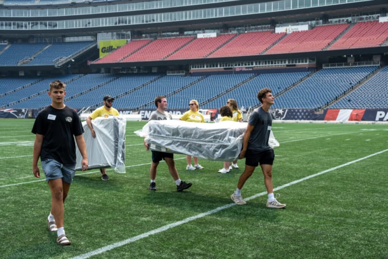 Group of young people carrying large plastic-wrapped items across a green football field with empty stadium seats in the background.