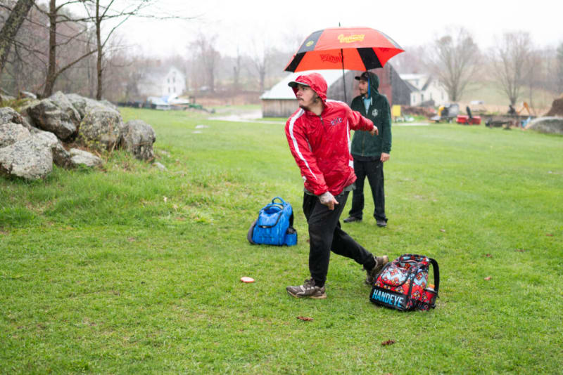 Person in red jacket and black pants throwing a disc in a grassy field, with another person holding a red and black umbrella nearby.