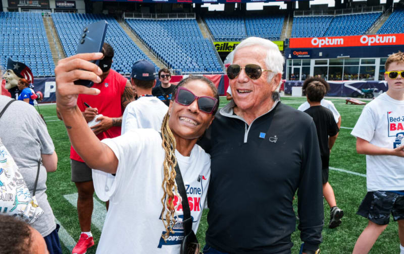 Two people taking a selfie on a football field with empty stadium seats in the background, one wearing a white t-shirt with a black strap and the other in a black quarter-zip pullover.