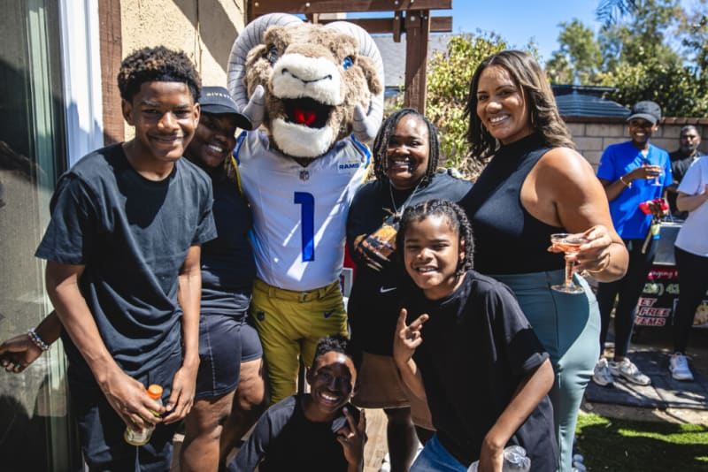 Group of people posing outdoors with a sports mascot wearing a white and gold Rams football uniform, some holding drinks, in a sunny backyard setting.