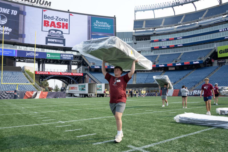 Man carrying a mattress wrapped in plastic on a football field during the Bed-Zone Bash event at a stadium with visible sponsor banners including Boston Scientific, jetBlue, and Toyota.