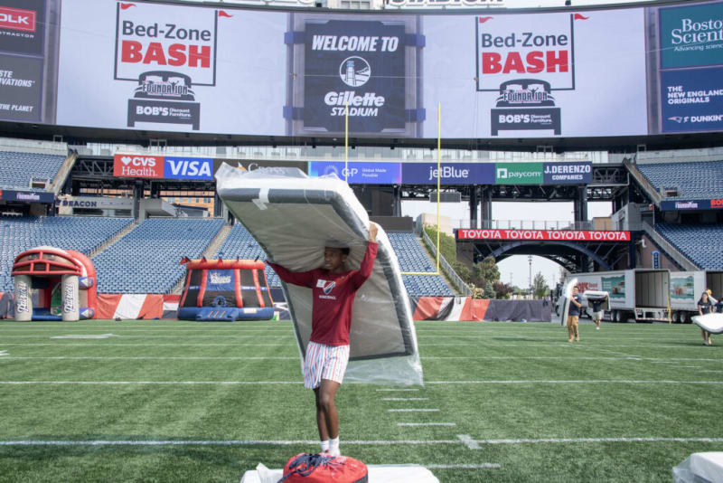 A person on a football field at Gillette Stadium holding a large mattress above their head. The stadium scoreboard displays "WELCOME TO Gillette STADIUM" and advertisements for Bed-Zone BASH, Boston Scientific, CVS Health, Visa, jetBlue, and Toyota.