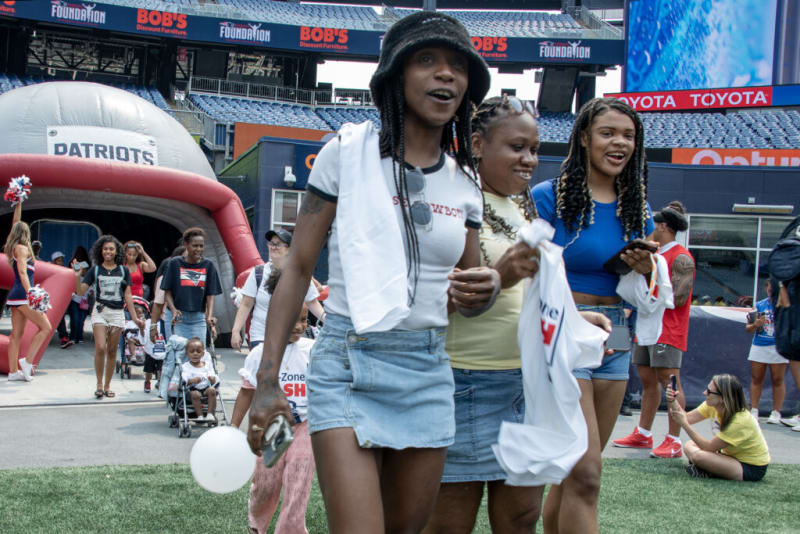 Three women in casual summer outfits, including denim skirts and shorts, holding white towels and balloons at a Patriots event in a stadium.
