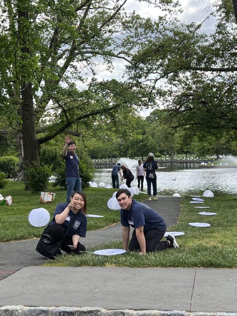 Group of people in navy blue shirts gathered near a pond with floating white lanterns, surrounded by green trees and a paved pathway.