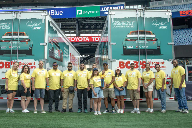 Group of people in yellow shirts standing in front of two green Bob’s Discount Furniture trucks with Toyota racing car graphics and text "Proud partner of Denny Hamlin & Joe Gibbs Racing."