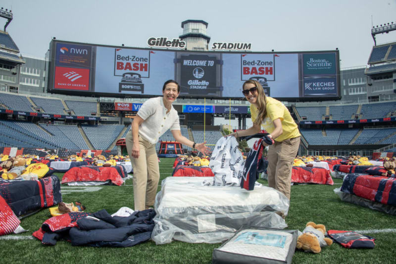 Two people setting up white and navy bedding with Patriots logos on a field at Gillette Stadium, surrounded by multiple beds with red, white, and navy blankets and stuffed bears, under a large scoreboard displaying "WELCOME TO Gillette STADIUM" and "Bed-Zone BASH."