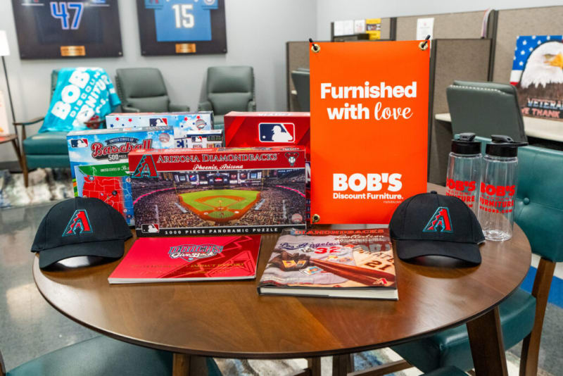 A round wooden table displaying Arizona Diamondbacks baseball merchandise including two black caps with the team logo, a 1000-piece panoramic puzzle, baseball-themed books, two clear water bottles with red Bob's Discount Furniture logos, and an orange sign reading "Furnished with love BOB'S Discount Furniture." The setting appears to be a casual office or showroom with chairs and sports memorabilia in the background.