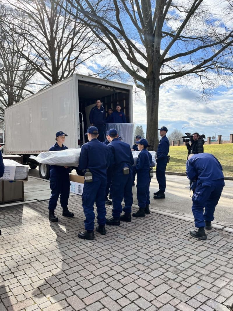 Group of uniformed personnel unloading large wrapped items from a white delivery truck on a paved outdoor area with leafless trees and blue sky.