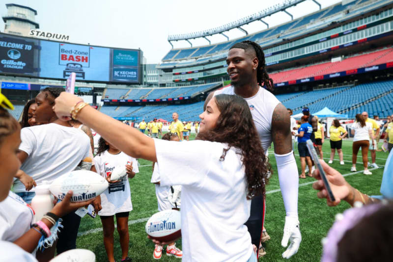 Fans taking selfies with a football player on the field at Gillette Stadium during the Bed-Zone Bash event, holding footballs and wearing event shirts.