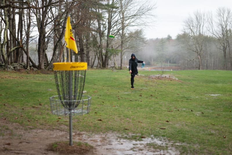Disc golf basket with yellow top and flag labeled "Discatcher" in a grassy park. A person in black clothing throws a green disc towards the basket.