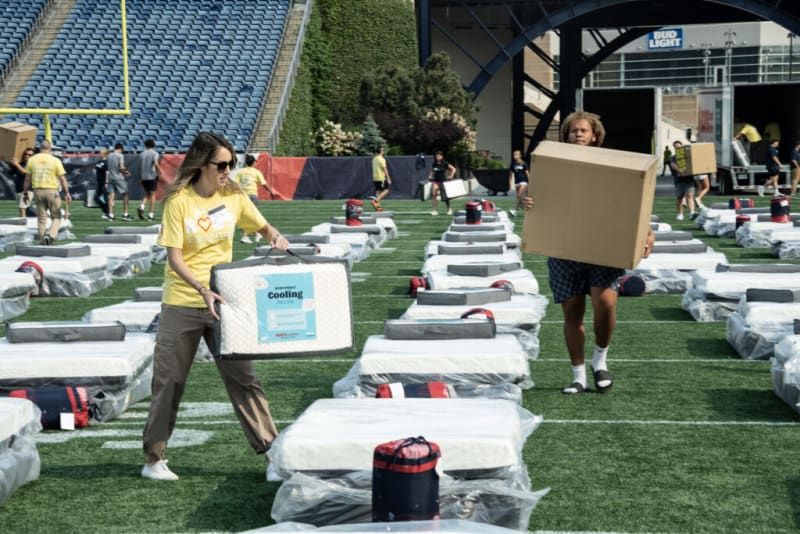 Volunteers wearing yellow shirts setting up a stadium field with wrapped mattresses and carrying cooling pillows and cardboard boxes for a relief or shelter event.