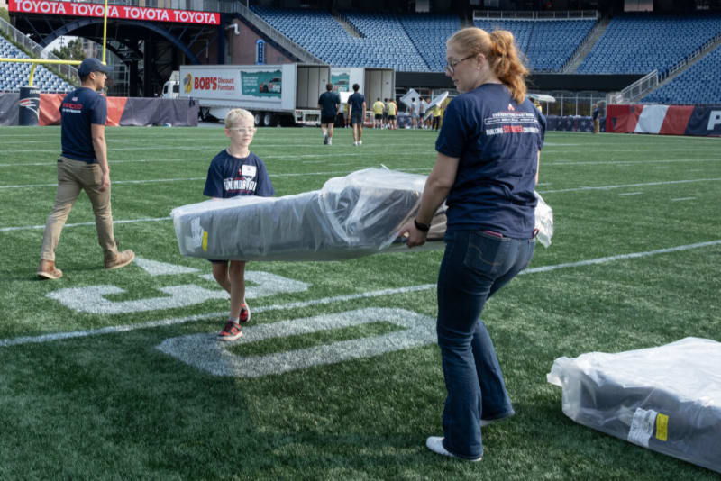 Two people carrying a large, wrapped mattress on a green football field with stadium seating and a truck labeled "BOB'S Discount Furniture" in the background. Both wear navy blue shirts with white and red text, indicating participation in a community or volunteer event. The scene suggests mattress delivery or setup at an outdoor event.