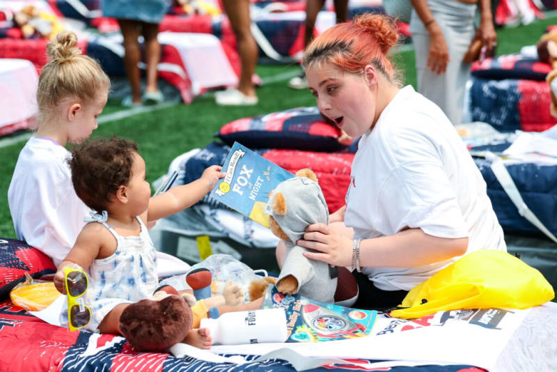 A toddler holding a book titled "I Can Read! FOX AT NIGHT" and yellow sunglasses, sitting on a red, white, and blue patterned blanket with a stuffed bear toy and an adult wearing a white shirt nearby.