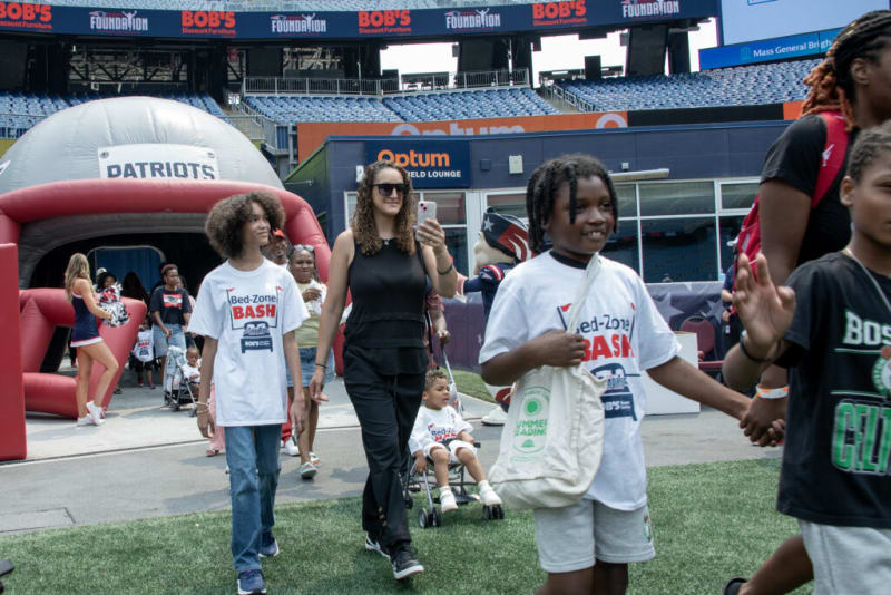 Group of people walking on a football field near a Patriots inflatable tunnel, wearing Bed-Zone Bash event t-shirts at a stadium event.