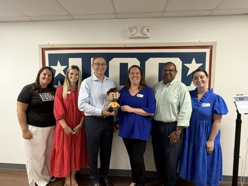 Group of six adults standing in front of a USO sign, with one person holding a small doll. The group wears business casual and casual attire, including a black USO t-shirt, red and blue dresses, and button-up shirts. The setting suggests a community or support event at a USO center.