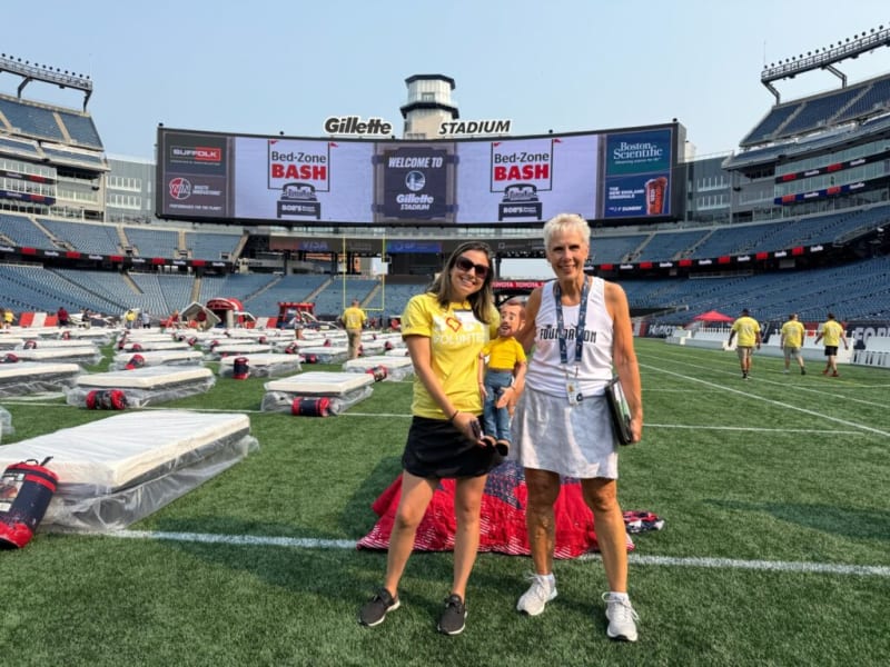 Volunteers standing on the field at Gillette Stadium with rows of cots set up for Bed-Zone Bash event, large stadium screen displaying event name and sponsors.