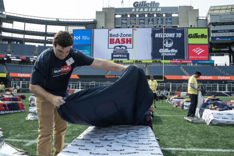 Man setting up a bed on a football field at Gillette Stadium for the Bed-Zone Bash event, with banners displaying "WELCOME TO Gillette STADIUM," "Bed-Zone BASH," and sponsor logos including Gillette and Bank of America.
