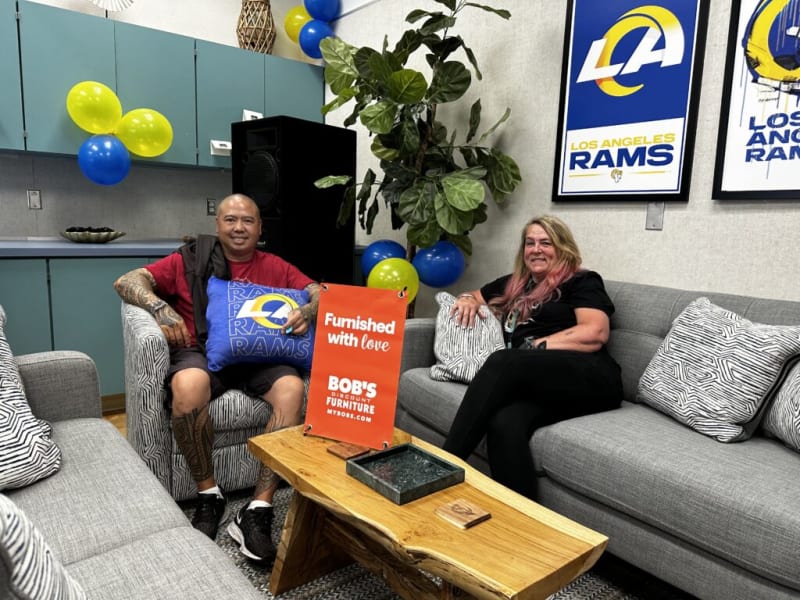 Two people sitting on patterned gray and white couches in a room decorated with Los Angeles Rams pillows, balloons, and framed Rams posters. A wooden coffee table with a black tray and a sign reading "Furnished with love BOB'S DISCOUNT FURNITURE MYBOBS.COM" is in the center. The setting suggests a cozy, sports-themed lounge or waiting area.