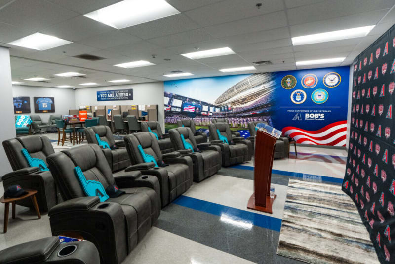 A modern lounge area with gray recliner chairs featuring blue foam fingers, a wooden podium, and a backdrop with military emblems and text: "THANK YOU FOR YOUR SERVICE * YOU ARE A HERO * TODAY, TOMORROW, AND ALWAYS" and "PROUD SUPPORTERS OF OUR VETERANS BOB'S DISCOUNT FURNITURE." The space includes sports memorabilia and is designed for veteran appreciation events.