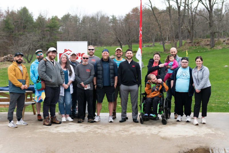 Group of people standing outdoors on a paved area with grass and trees in the background, some wearing casual jackets and sneakers, one person in a wheelchair, and a red and white flag behind them.