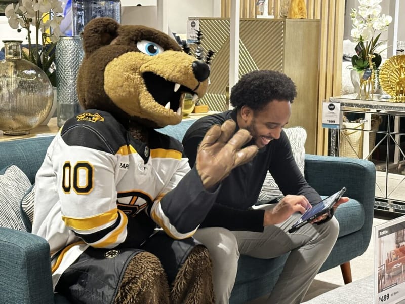 Person sitting on a blue sofa using a tablet next to a Boston Bruins mascot in a white and yellow hockey jersey with number 00, in a modern furniture store.