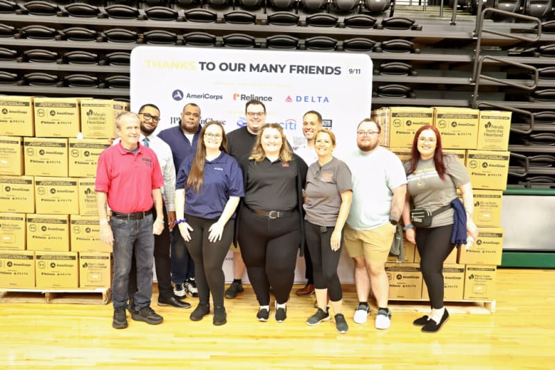 Group of eight people standing in front of a banner that reads "THANKS TO OUR MANY FRIENDS 9/11" with logos of AmeriCorps, Reliance, and Delta, surrounded by stacks of yellow boxes labeled "Meals from the Heartland."