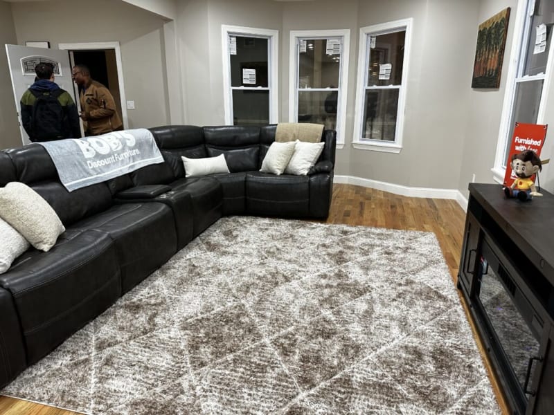 Black leather sectional sofa with beige and white cushions, a gray patterned area rug on hardwood floor, and a dark wood TV stand with a small plush toy. A gray towel with "BOB'S Discount Furniture" text draped over the sofa. Three white-framed windows and neutral beige walls in the background.