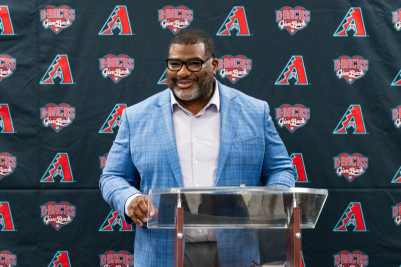 Man in blue plaid blazer and light purple shirt standing behind a clear podium with Arizona Diamondbacks "D-Backs Give Back" logo backdrop.
