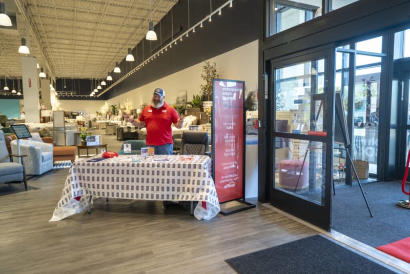 Retail store interior with a promotional table covered in a white and blue checkered tablecloth, displaying brochures and small items. Next to the table is a tall digital sign with red background text promoting convenient payment options: "convenient ways to pay," "No money down," "No credit needed," "Special financing," "Low monthly payments," and "4 interest-free payments with Affirm." The setting includes modern furniture and large glass doors.