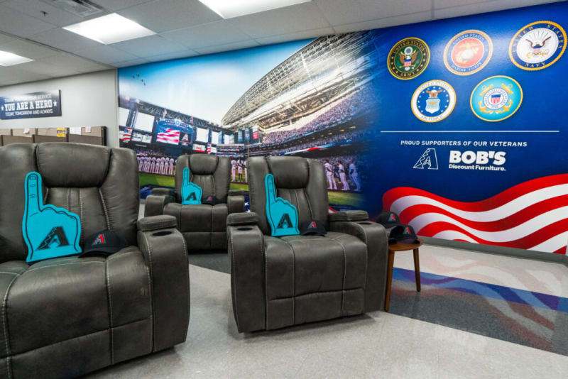 Modern lounge area with black leather recliners featuring blue foam finger pillows and Arizona Diamondbacks caps, set against a patriotic mural with U.S. military emblems and text: "THANK YOU FOR YOUR SERVICE YOU ARE A HERO TODAY, TOMORROW AND ALWAYS" and "PROUD SUPPORTERS OF OUR VETERANS BOB'S Discount Furniture."