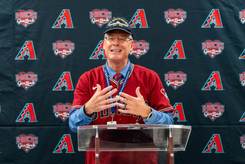 Man wearing a red Arizona Diamondbacks jersey and a blue shirt with a striped tie, speaking at a podium with a backdrop featuring "D-Backs Give Back" and Diamondbacks logos.