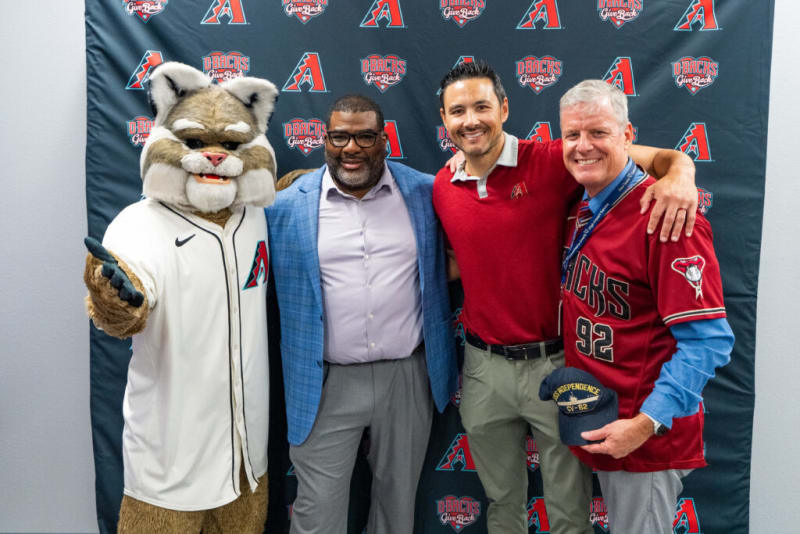 A person in a white Arizona Diamondbacks mascot costume stands next to two men, one wearing a red polo with an Arizona Diamondbacks logo and the other in a red Diamondbacks jersey holding a navy blue cap with "USS INDEPENDENCE CV-62" embroidered. The backdrop features repeating Arizona Diamondbacks logos and "D-Backs Give Back" text.