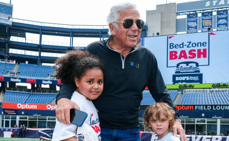 Man in a black quarter-zip pullover and blue jeans with two children wearing white event t-shirts at a stadium. A large screen behind them displays "Bed-Zone BASH" with logos for Foundation and Bob's Discount Furniture, indicating a promotional or charity event. Stadium seating and banners for Optum and Gillette are visible in the background.