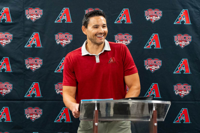 Man wearing a red Arizona Diamondbacks polo shirt standing behind a clear podium with a D-backs Give Back logo backdrop.