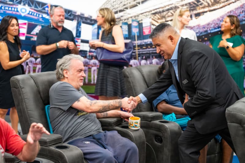 A man in a black suit shakes hands with a seated man wearing a gray t-shirt and blue pants in a stadium lounge area with leather recliners and a baseball field backdrop.