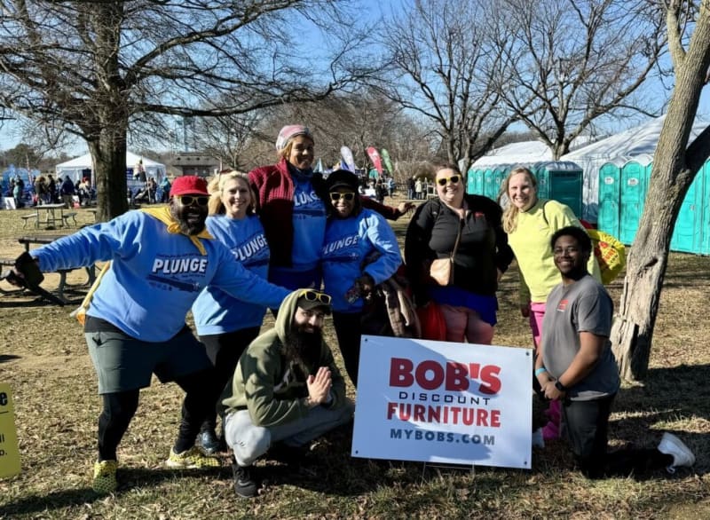 Group of people outdoors at a charity event holding a sign that reads "BOB'S DISCOUNT FURNITURE MYBOBS.COM" with portable restrooms and tents in the background.