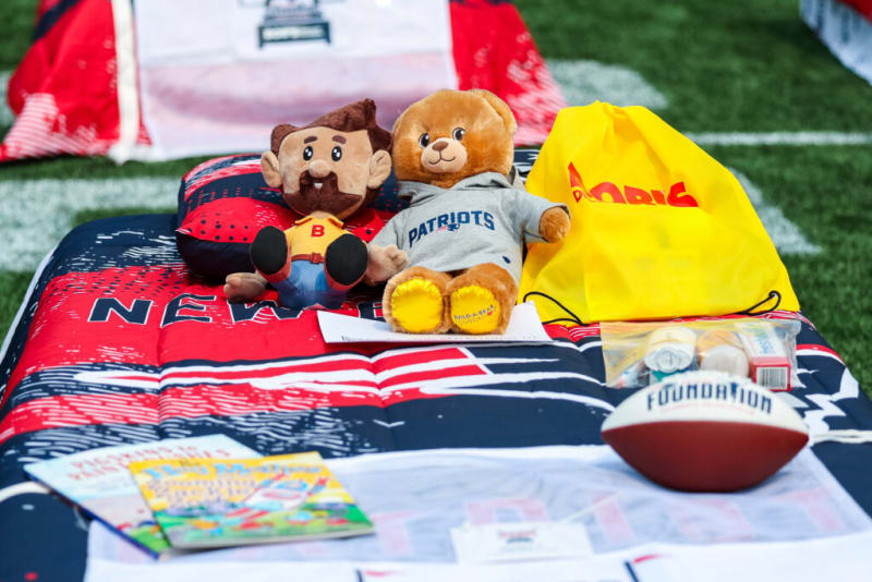 A New England Patriots-themed bedding set on artificial turf with plush toys, including a bear wearing a Patriots hoodie, a football labeled "Foundation," a yellow bag, and children's books.