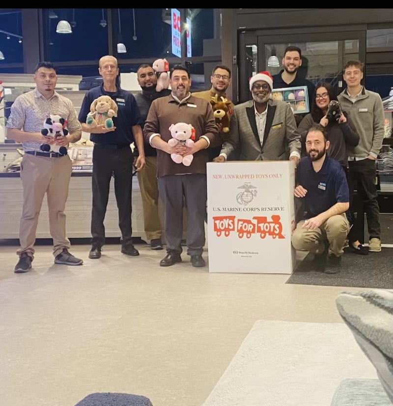 Group of men standing indoors holding stuffed toys next to a large white donation box labeled "NEW, UNWRAPPED TOYS ONLY U.S. MARINE CORPS RESERVE TOYS FOR TOTS" promoting toy donations.