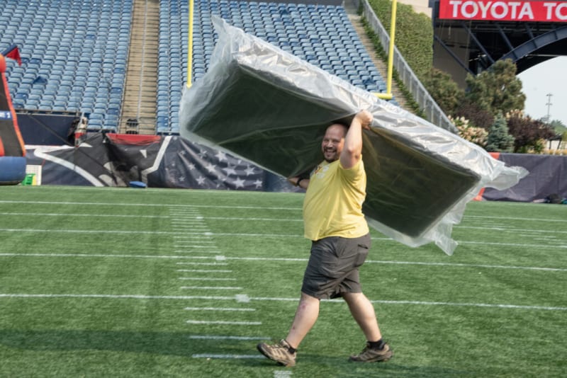 Man carrying a large, plastic-wrapped mattress or cushion across a green football field with empty blue stadium seats and a Toyota sign in the background.
