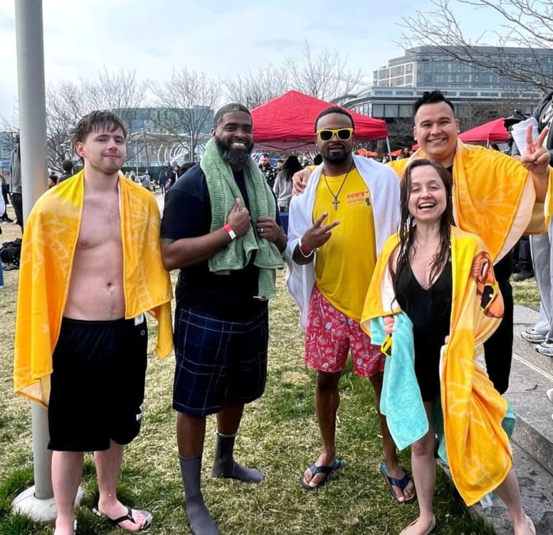 Group of five people outdoors wearing yellow and white towels, casual swimwear, and sandals, standing on grass near tents and buildings.