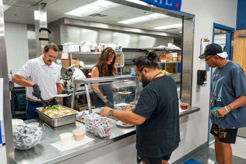 People serving and receiving food at a cafeteria-style counter with trays of salad, pizza, and condiments in a commercial kitchen setting.