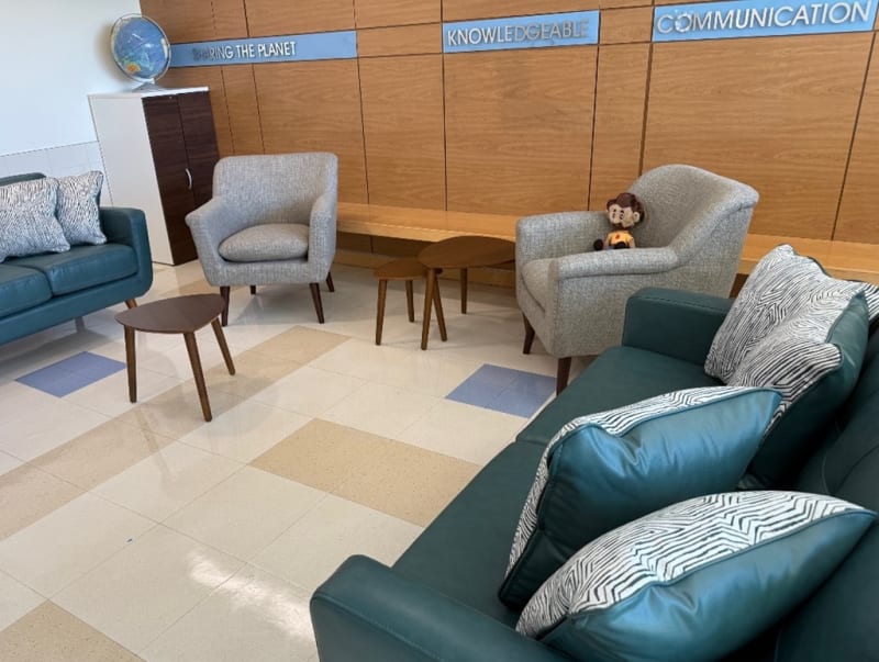Modern office lounge area with teal leather sofas, gray fabric armchairs, patterned cushions, wooden side tables, and wall signs reading "SHARING THE PLANET," "KNOWLEDGEABLE," and "COMMUNICATION."