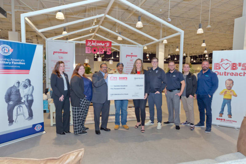 Group of people standing inside a Bob's Furniture store holding a large check for $50,000 made out to Operation Homefront, promoting Bob's Outreach program.