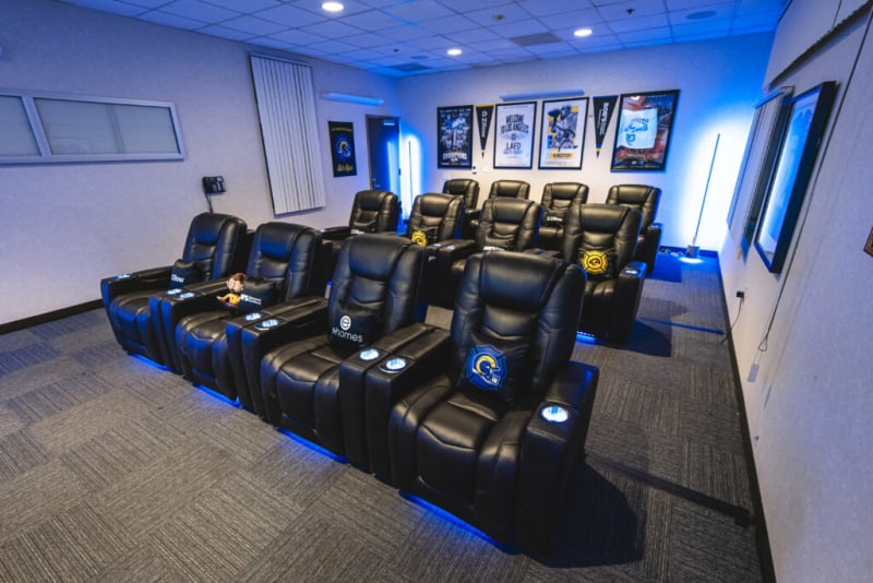 A modern home theater room with two rows of black leather reclining chairs featuring blue LED lighting underneath. The chairs have branded pillows and cup holders. The room has framed sports posters on the back wall and ambient blue lighting, creating a cozy entertainment space.