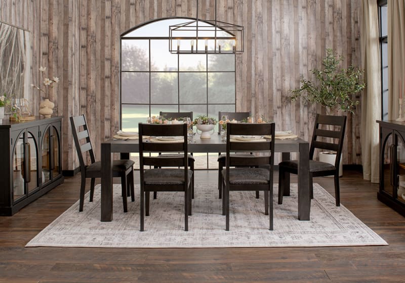 Dining room with dark wood table and six matching ladder-back chairs on a light patterned rug, featuring rustic wood panel walls and large window.