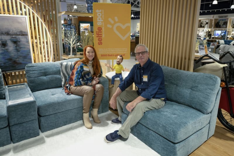 Two people sitting on modern blue fabric sofas in a retail furniture store, with a yellow sign behind them reading "selfie spot" and "$500 Bob's gift card!"