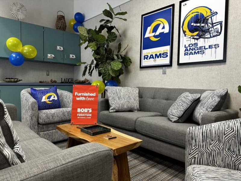 Modern living room setup featuring gray fabric sofas and armchairs with black-and-white patterned cushions, wooden coffee table, and Los Angeles Rams framed wall art. Bright yellow, blue, and green balloons add color, with a red sign reading "Furnished with love BOB'S DISCOUNT FURNITURE MYBOBS.COM" on the table. The space combines comfort and team spirit decor.