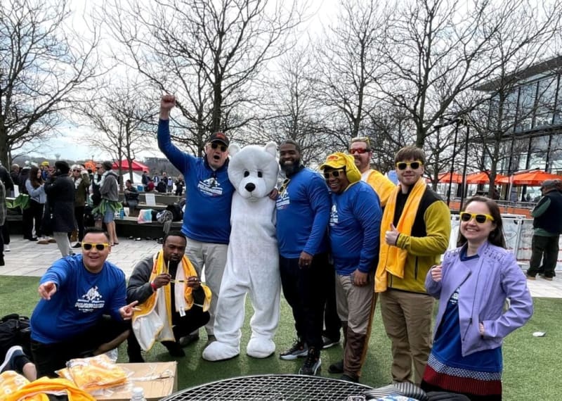 Group of people wearing blue Polar Plunge shirts and yellow towels posing outdoors with a person in a white polar bear costume on a grassy area.
