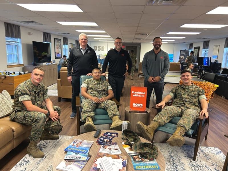 A group of military personnel and staff in a USO lounge with wood flooring and modern furniture. Two soldiers in camouflage uniforms sit in teal armchairs, one on a brown leather sofa, and three staff in black and gray tops stand behind them. A small orange sign on a round wooden table reads "Furnished with love BOB'S Discount Furniture." The room features large windows, a foosball table, and a TV, creating a comfortable recreational space for service members.